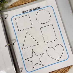 May include: A white worksheet in a binder with the text "Trace the Shapes" and outlines of a square, circle, rectangle, triangle, star, and heart. The shapes are outlined with dashed lines for tracing. The worksheet is in a clear plastic sleeve.