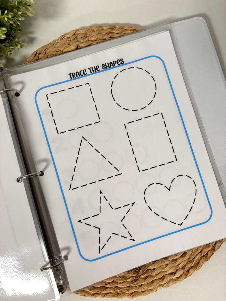 May include: A white worksheet in a binder with the text "Trace the Shapes" and outlines of a square, circle, rectangle, triangle, star, and heart. The shapes are outlined with dashed lines for tracing. The worksheet is in a clear plastic sleeve.