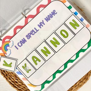 May include: A binder with a colorful cover featuring the text "I CAN SPELL MY NAME." The spelling activity includes letter tiles to spell the name "KANNON." A rainbow and cloud graphic is in the corner. The binder is open on a woven basket.