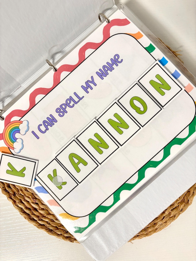 May include: A binder with a colorful cover featuring the text "I CAN SPELL MY NAME." The spelling activity includes letter tiles to spell the name "KANNON." A rainbow and cloud graphic is in the corner. The binder is open on a woven basket.