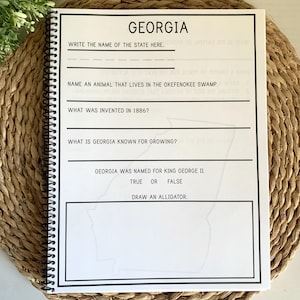 Puede incluir: Un cuaderno con espiral con una cubierta en blanco y negro con la palabra "GEORGIA" y preguntas para completar sobre el estado. El cuaderno est&aacute; abierto sobre una estera tejida, con una planta verde en el fondo.