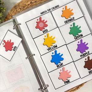 May include: A binder with pages featuring color matching activities. Each page displays colorful paint splatters labeled with color names like red, orange, yellow, green, blue, purple, pink, and brown. The text "MATCH THE COLORS!" is at the top.
