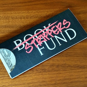 Op de afbeelding: Een zwarte kaart met de woorden "BOOK FUND" in het wit, met "STRIPPERS" in het roze doorgestreept. Een dollarbiljet is gedeeltelijk zichtbaar in de zak van de kaart. De kaart ligt op een houten oppervlak.