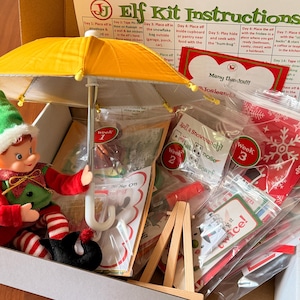May include: A holiday elf kit in a cardboard box containing a small elf doll holding a yellow umbrella, a printed instruction sheet titled Elf Kit Instructions, and several clear plastic bags filled with various craft supplies and holiday-themed items.