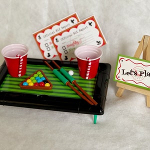 May include: A miniature pool table with green felt, red and white cups, and wooden cues. The table has a black frame and green legs. A sign on a small easel reads "Let's Play!"