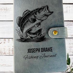May include: Gray fishing journal with a black and white illustration of a largemouth bass leaping out of the water. The journal has the text "JOSEPH DRAKE Fishing Journal" and a tassel.