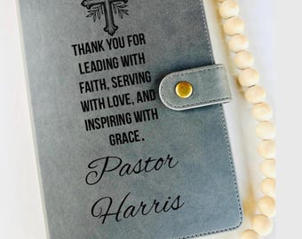 May include: Grey leather-effect journal featuring a cross emblem and the inscription "THANK YOU FOR LEADING WITH FAITH, SERVING WITH LOVE, AND INSPIRING WITH GRACE." The name "Pastor Harris" is below. A wooden bead strand is on the right.