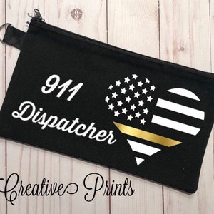 May include: Black zippered pouch with a white heart design featuring an American flag and a gold stripe. The text "911 Dispatcher" is printed in white on the pouch.