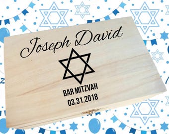 May include: A light-colored wooden box with the name "Joseph David" in elegant script, a black Star of David, and the text "Bar Mitzvah 03.31.2018". The box is set against a background of blue and white Star of David and balloon patterns.