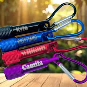 May include: A collection of personalized LED flashlights in black, blue, red, and purple, each with a silver carabiner clip. The flashlights are engraved with names: Kyle, Cristiano, William, and Camila. The flashlights are compact and portable.