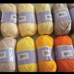 May include: A collection of yarn skeins in various colors, including cream, yellow, orange, and green. Each skein is wrapped with a purple label. The yarn is tightly wound, suggesting it's ready for knitting or crocheting projects.