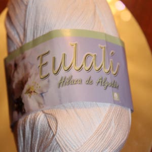 May include: A ball of light beige cotton yarn with a label that reads "Eulali Hilaza de Algod&oacute;n". The yarn is tightly wound, and the label features floral imagery. Ideal for knitting and crochet projects.