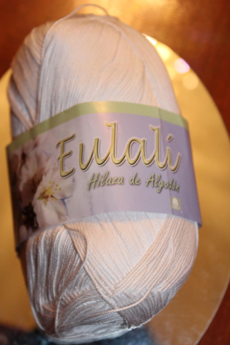 May include: A ball of light beige cotton yarn with a label that reads "Eulali Hilaza de Algod&oacute;n". The yarn is tightly wound, and the label features floral imagery. Ideal for knitting and crochet projects.