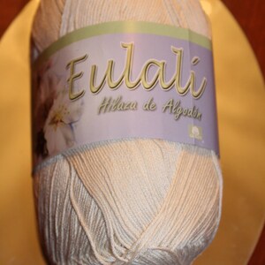 May include: A ball of light beige yarn with a label that reads "Eulali Hilaza de Algod&oacute;n". The label has a floral design and is wrapped around the center of the yarn. The yarn is on a reflective surface.