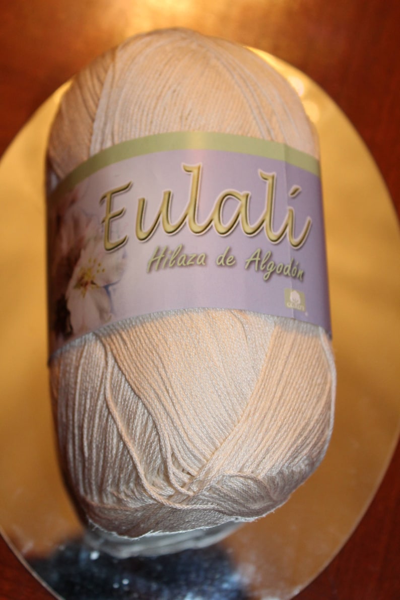 May include: A ball of light beige yarn with a label that reads "Eulali Hilaza de Algod&oacute;n". The label has a floral design and is wrapped around the center of the yarn. The yarn is on a reflective surface.
