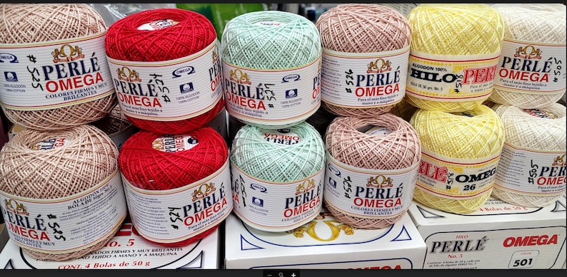 May include: A collection of Perle Omega cotton yarn balls in various colors, including red, beige, green, pink, yellow, and white. Each ball is labeled with the brand name and product information. Ideal for crochet and knitting projects.