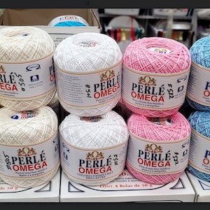 May include: A display of colorful yarn balls, including yellow, white, pink, and blue. Each ball is labeled "PERLE OMEGA" and contains 50 grams of 100% cotton yarn. The yarn is arranged on a shelf, ready for crafting.