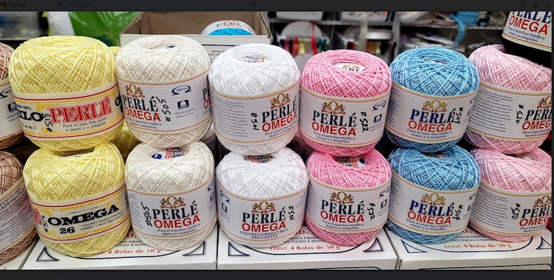May include: A display of colorful yarn balls, including yellow, white, pink, and blue. Each ball is labeled "PERLE OMEGA" and contains 50 grams of 100% cotton yarn. The yarn is arranged on a shelf, ready for crafting.