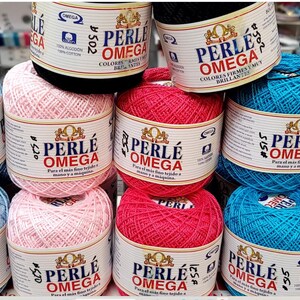 May include: A display of colorful Perle Omega cotton yarn balls in various shades, including pink, blue, red, and black. Each ball is labeled with the brand name and product details. The yarn is 100% cotton.