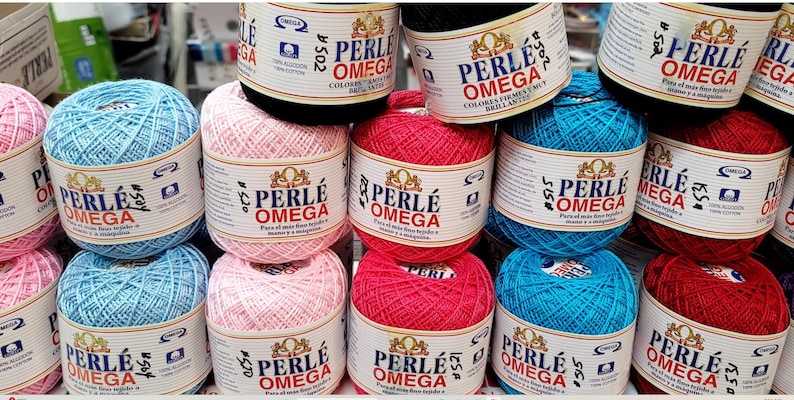 May include: A display of colorful Perle Omega cotton yarn balls in various shades, including pink, blue, red, and black. Each ball is labeled with the brand name and product details. The yarn is 100% cotton.