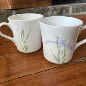 May include: Two white ceramic mugs with floral designs. One mug features a blue iris with green leaves and the other mug has a green stem with a blue flower bud.