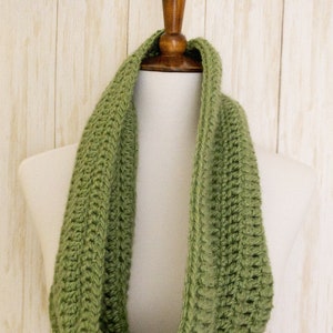 May include: A green crocheted infinity scarf, displayed on a white mannequin against a woodgrain background.