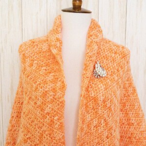 May include: An orange and white crocheted shawl with a decorative brooch. The shawl is draped over a mannequin, showcasing its open front design and scalloped edges. The brooch is silver with clear stones.
