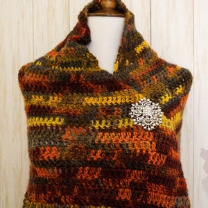 May include: A brown, orange, and yellow crocheted shawl with a sparkly brooch pinned to it. The shawl is draped over a white mannequin.