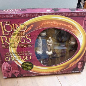 May include: A rectangular box for The Lord of the Rings: The Two Towers chess set with an antique ivory and Chinese lacquer finish. The packaging features a large circular window displaying sculpted chess pieces and character imagery.