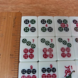 May include: A set of white Mahjong tiles featuring colorful engraved symbols and numbers, arranged in a grid next to a wooden ruler showing measurements in inches, with visible tiles displaying the numbers 4, 5, 7, 9, and 1.
