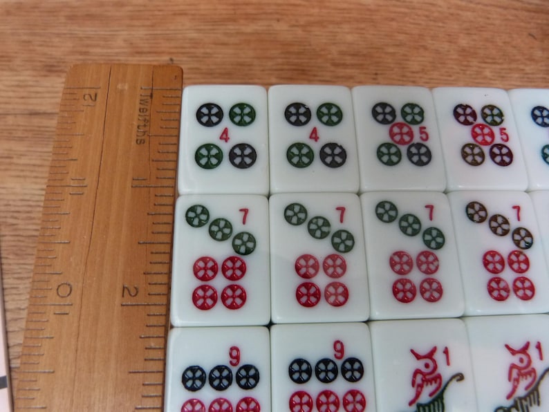 May include: A set of white Mahjong tiles featuring colorful engraved symbols and numbers, arranged in a grid next to a wooden ruler showing measurements in inches, with visible tiles displaying the numbers 4, 5, 7, 9, and 1.
