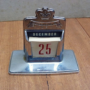 May include: A silver-toned metal perpetual desk calendar featuring a crown emblem at the top with the text Elizabeth R 1953 engraved below. The calendar displays the month of December in black text and the day 25 in large red numerals on white pages.