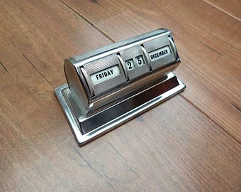 May include: A silver-toned, vintage perpetual desk calendar displaying "FRIDAY 25 DECEMBER." The calendar has a rectangular base and a rotating mechanism to change the date and day. The calendar is made of metal and sits on a wooden surface.