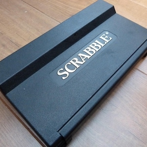 May include: A black Scrabble game box with the word "SCRABBLE" in silver lettering. The box is rectangular with a raised section on the top. The game is set on a wooden surface.