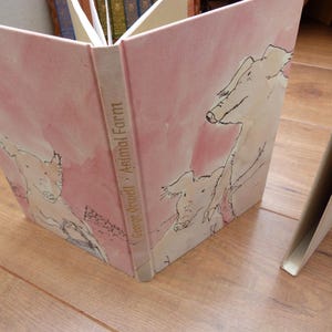 May include: A pink book with a watercolour design featuring pigs. The book's spine has the title "George Orwell - Animal Farm" in gold lettering.