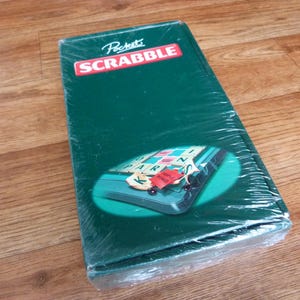 May include: A green Pocket Scrabble game in its original packaging. The game box has the word "Scrabble" in a red rectangle. The game is sealed in clear plastic wrap and sits on a wooden surface.