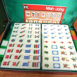 May include: A Mah-Jong game set in a green zippered case containing white tiles with colorful symbols, numbers, and floral designs. The set includes a red instruction card labeled KTG Know the Game Mah-Jong, stacks of green and yellow chips, and dice.