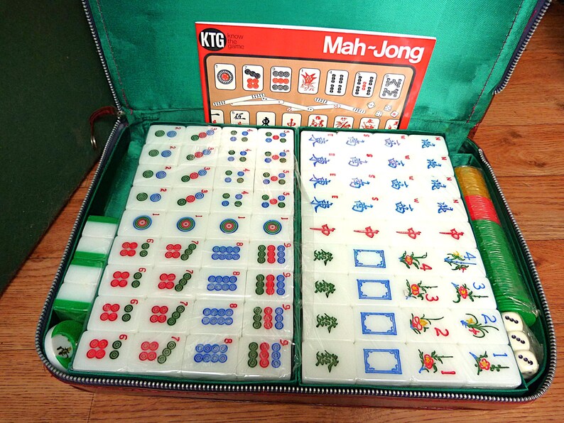 May include: A Mah-Jong game set in a green zippered case containing white tiles with colorful symbols, numbers, and floral designs. The set includes a red instruction card labeled KTG Know the Game Mah-Jong, stacks of green and yellow chips, and dice.