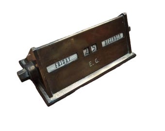May include: A vintage brass desk calendar showing "Friday, December 25" and the initials "E.G." The calendar has a rectangular base and a rotating mechanism. The numbers and letters are in a classic font. The calendar is made of brass.