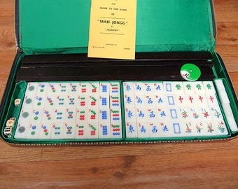 May include: A Mah-Jongg game set in a zippered carrying case with a green interior. The set includes white tiles with red, blue, and green markings, dice, and a yellow instruction booklet titled GUIDE TO THE GAME OF MAH-JONGG BY JACKPOT.
