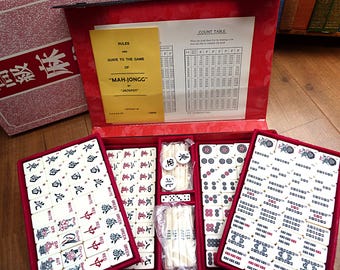 Puede incluir: Un juego de Mah-Jong en un estuche rojo con fichas blancas que presentan símbolos en negro, rojo y verde. El conjunto incluye un folleto de instrucciones amarillo titulado Rules and Guide to the Game of Mah-Jongg by Jackpot y una tabla de conteo.