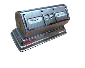 May include: A vintage silver desk calendar showing "FRIDAY 25 DECEMBER." The calendar has a rectangular base and rotating panels. The calendar is made of metal with a textured design. The measurements are in centimetres.