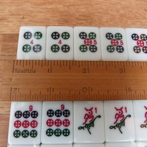 May include: A set of white rectangular Mahjong tiles with colorful engraved symbols and numbers, arranged in two rows. A wooden ruler is placed across the tiles, showing a measurement of approximately 4.5 inches across the top row of tiles.