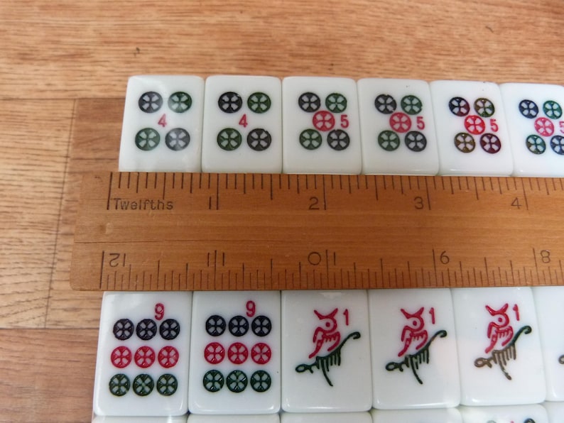 May include: A set of white rectangular Mahjong tiles with colorful engraved symbols and numbers, arranged in two rows. A wooden ruler is placed across the tiles, showing a measurement of approximately 4.5 inches across the top row of tiles.
