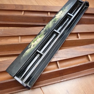 May include: Wooden Scrabble tile holders and a black tile rack with a landscape design. The wooden holders are a medium brown colour, and the black rack has a painted scene with white and green accents. The items are on a wooden surface.