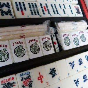Peut inclure: Un jeu de Mahjong compos&eacute; de tuiles blanches avec des symboles grav&eacute;s en noir, rouge et bleu, incluant des tuiles Joker, des tuiles saisonni&egrave;res marqu&eacute;es Winter et Chrys, et des tuiles directionnelles, rang&eacute;es dans un plateau noir.