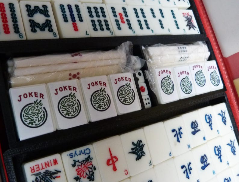 Peut inclure: Un jeu de Mahjong compos&eacute; de tuiles blanches avec des symboles grav&eacute;s en noir, rouge et bleu, incluant des tuiles Joker, des tuiles saisonni&egrave;res marqu&eacute;es Winter et Chrys, et des tuiles directionnelles, rang&eacute;es dans un plateau noir.