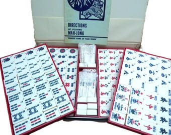 May include: A vintage Mah-Jong set with red trays and white tiles. The tiles feature various symbols and characters in red, green, and blue. The set includes a booklet titled "Directions of Playing Mah-Jong" and is stored in a white box.