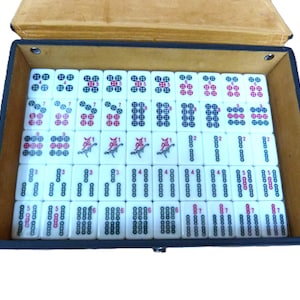 May include: A set of white rectangular mahjong tiles arranged in a black storage case with a tan interior. The tiles feature various black and red circular patterns, numerical markings, and bird illustrations, organized in neat rows and columns.