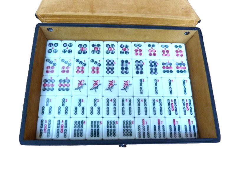 May include: A set of white rectangular mahjong tiles arranged in a black storage case with a tan interior. The tiles feature various black and red circular patterns, numerical markings, and bird illustrations, organized in neat rows and columns.